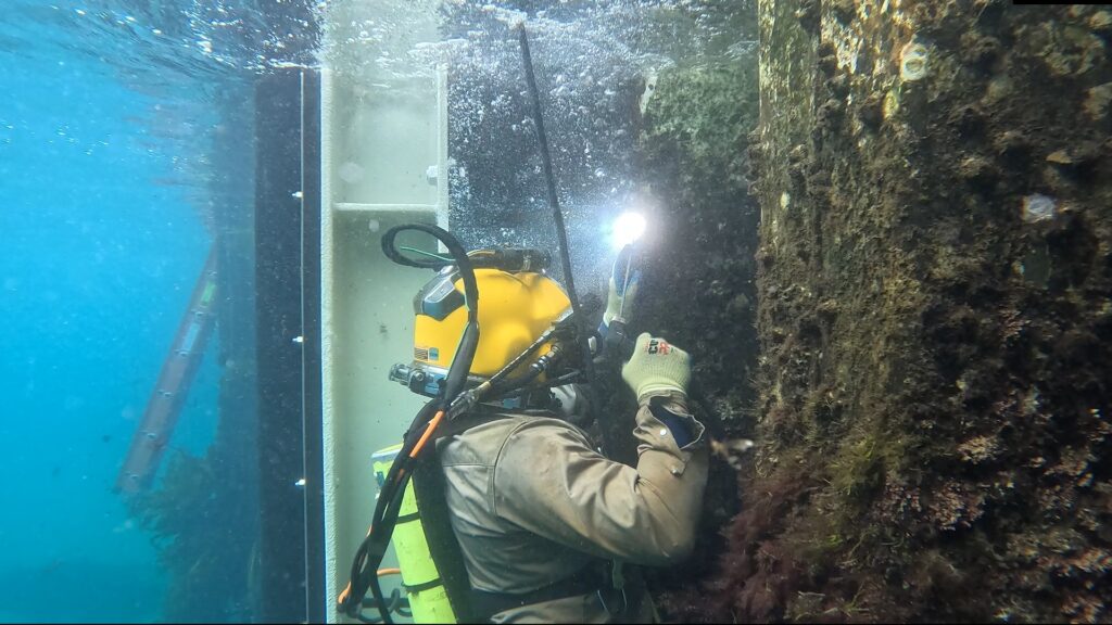 A diver inspects underwater structures with equipment and a flashlight.