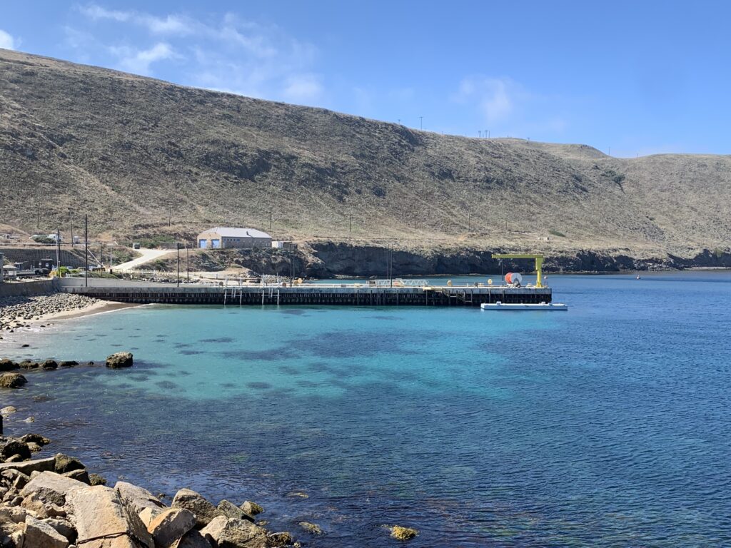 Clear turquoise water by a rocky coastline with a pier and buildings under a blue sky.